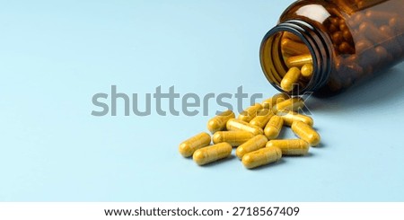 Amber supplement bottle tips over as yellow capsules rest on clean blue background with copy space for wellness advertising.