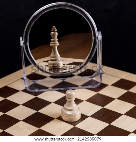 An ambitious chess pawn on a chessboard looks in the mirror and sees himself as a queen or king. The concept of confidence, ambition, self-belief,success,business.