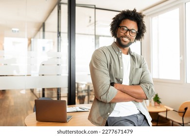 Ambitious highly-skilled handsome curly african-american male employee in glasses and casual wear leaned on office desk and looking at the camera with smile, businessman stands with arms crossed - Powered by Shutterstock