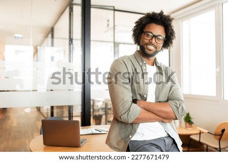 Ambitious highly-skilled handsome curly african-american male employee in glasses and casual wear leaned on office desk and looking at the camera with smile, businessman stands with arms crossed