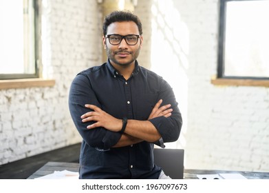 Ambitious hindu man in smart casual shirt stands with arms crossed in contemporary office space and looks at camera, portrait of purposeful indian businessman, ceo indoor. Diversity of office staff