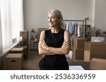 Ambitious mid aged lady warehouse or web store holder stand with arms crossed in confident pose look aside with smile enjoy perfect sales results. Happy small company owner proud with reaching success