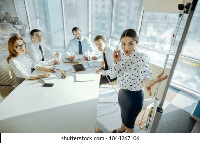 Ambitious Objectives. Charming Young Woman Pointing At The Whiteboard During The Meeting While Telling Her Team The Goals For Their New Project