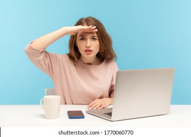 Ambitious Young Woman Employee Sitting At Workplace With Laptop And Looking Far Away With Hand Above Eyes, Searching With View, Exploring Distant Future. Indoor Studio Shot Isolated On Blue Background