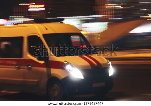 An ambulance car rides through the city at night\
amid glowing lights.