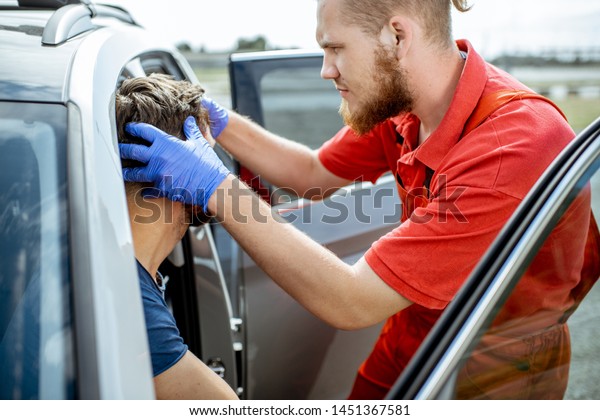 Ambulance worker examining facial injuries of\
a man sitting near the car after the road accident, providing\
emergency medical\
assistance