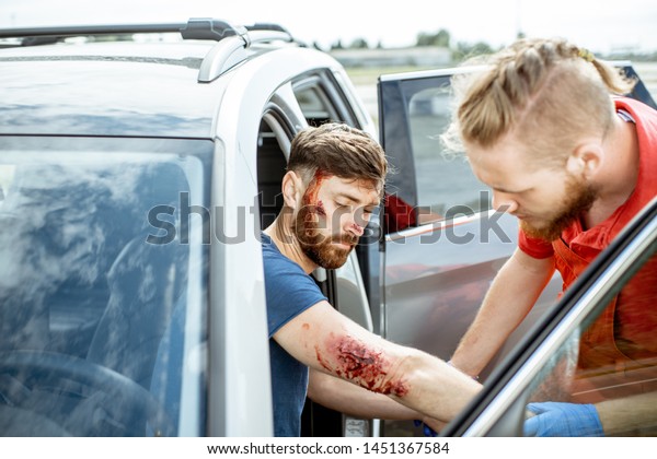 Ambulance worker examining injuries of a man\
sitting near the car after the road accident, providing emergency\
medical assistance