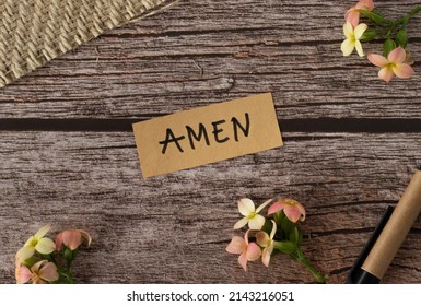 Amen, handwritten text with uppercase letters in English on wooden table with flowers and pen in vintage style. Top view, flay lay.