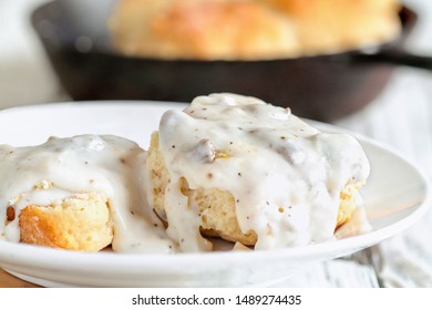 American biscuits from scratch covered with thick white sausage gravy. Selective focus with cast iron skillet / pan in the background over a white table.  - Shutterstock ID 1489274435