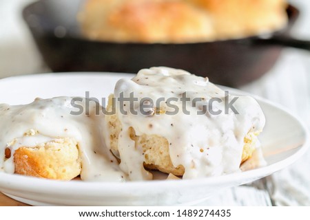 American biscuits from scratch covered with thick white sausage gravy. Selective focus with cast iron skillet / pan in the background over a white table. 