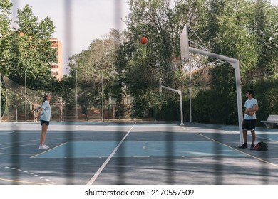 American Blonde Woman Shooting A Street Basketball Free Throw. Young Girl Scoring A Two-point Basket On A Blue Court While A Boy Who Is Her Rival Watches Her. Friendship And Sport Concept