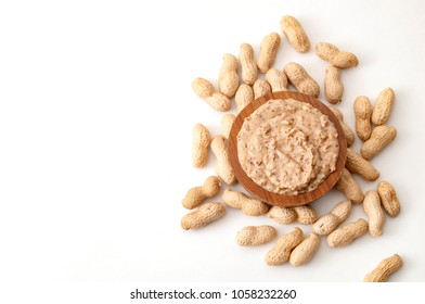 American Breakfast, Sweet Food And Snack High In Calories Concept With A Bowl Of Crunchy Peanut Butter Surrounded By Peanuts Still In Shell, Isolated On White Background With Copy Space