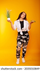 American Brunette Girl In One Hand Holding Grass, In The Other Burger Fast Food Junk Food On A Yellow Background In Isolation