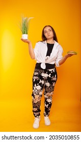 American Brunette Girl In One Hand Holding Grass, In The Other Burger Fast Food Junk Food On A Yellow Background In Isolation