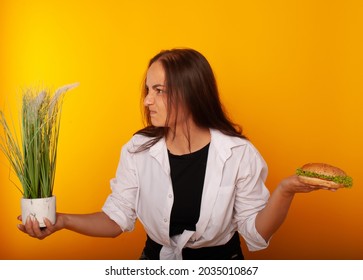 American Brunette Girl In One Hand Holding Grass, In The Other Burger Fast Food Junk Food On A Yellow Background In Isolation