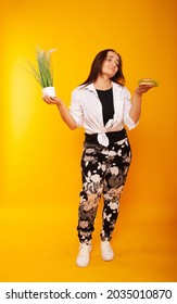 American Brunette Girl In One Hand Holding Grass, In The Other Burger Fast Food Junk Food On A Yellow Background In Isolation