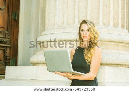American Business Woman with long blonde hair working in New York, wearing sleeveless black dress, standing by column on street, reading, working on laptop computer, thinking. Color filtered effect