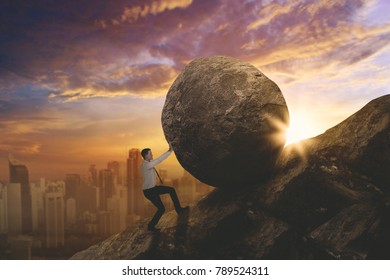 American businessman struggling to push a stone while climbing on the cliff. Shot at sunset - Shutterstock ID 789524311