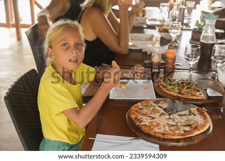 American curly blonde boy eating pizza in italian restaurant. Portrait of child with long blonde hair and a yellow T-shirt eats junk food in a cafe. Gastronomic trip in Italy.