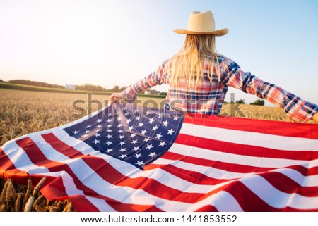 American female farmer in casual clothing with arms spread open holding USA flag in wheat field.