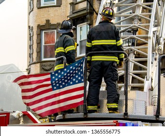 American Firefighters On Fire Ladder Truck Turntable With Flag Blowing In The Wind