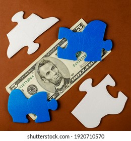 The American Five Dollar Bill Is Hidden Under The Puzzle Pieces. Blue And White Puzzles With Money On A Brown Background. Top View, Flat Lay.