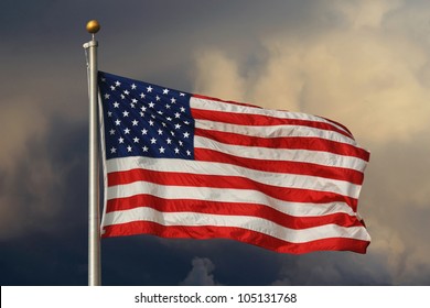 American flag in foreground, waving and brightly lit with ominous clouds in the background. / Old Glory Aglow / Brightly lit stars and stripes.