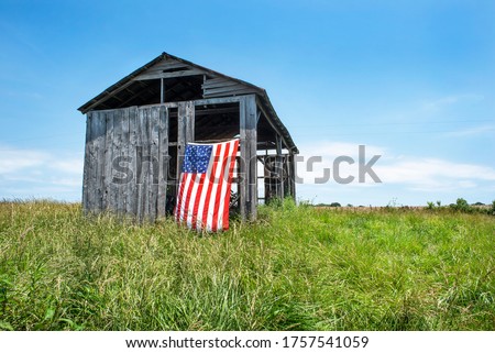 American flag hanging outdoors on side of old gray wooden barn on grass hill in countryside. Pride, patriotism, holidays, symbol, icon, independent, heartland, corn belt, farmers, country, vote, voter