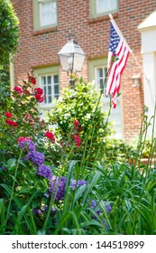 An American Flag On A Nice Brick House With A Lush Spring Garden In Front