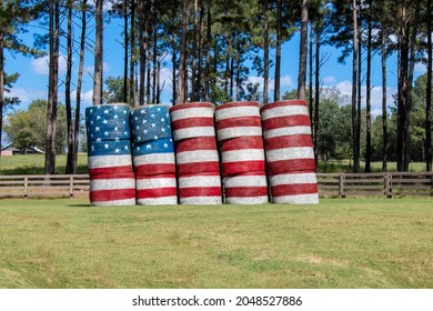 An American flag is painted across a stack of large round hay bales in a farm field against a backdrop of pine trees and blue sky.