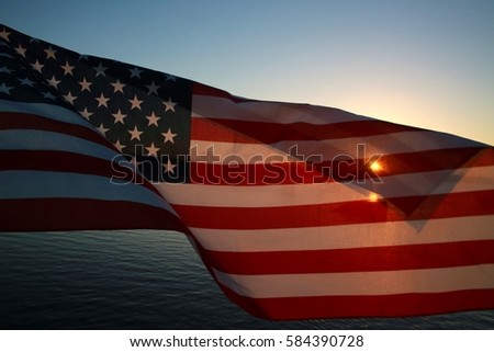 American Flag Stars and Stripes Flapping in the Breeze with Yellow Orange Setting Sun Half-Tansparent Gleaming Through, Water Below and Blue Sky Above, Quiet Waters Park Lake, Deerfield Beach, Florida