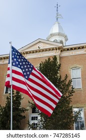 An American flag waves in the wind in front of a nondescript Victorian era courthouse.  The cupola, weathervane and gingerbread trim on the courthouse give a sense of history.