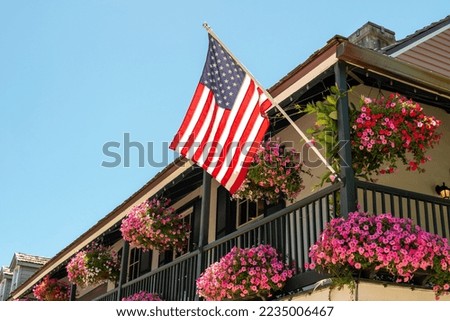 American flags hang off a second floor balcony of a historic building. The railings have baskets of pink and purple flowers hanging. The sky is blue and there are multiple windows in the old building.