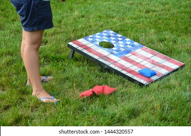 American flats corn hole game board in the green grass with bean bags and person’s legs with blue flip flops on