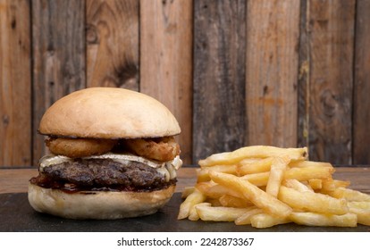 American food. Closeup view of a delicious rustic burger with meat, fried onion rings, brie cheese and sweet chili sauce, with french fries, in a black dish with a wooden background. - Powered by Shutterstock