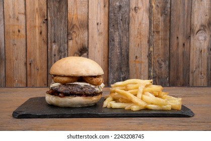 American food. Closeup view of a delicious rustic burger with meat, fried onion rings, brie cheese and sweet chili sauce, with french fries, in a black dish with a wooden background. - Powered by Shutterstock