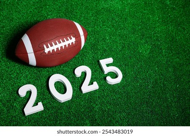 American football ball next to 2025 number whose figures are made of white painted wood isolated over green grass seen from above.