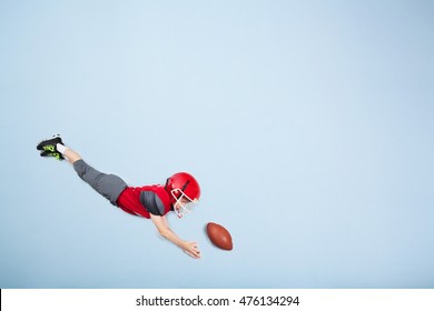 American Football Kid Wearing Full Pads And Helmet Is Diving For The Catch. Blue Background With Copy Space