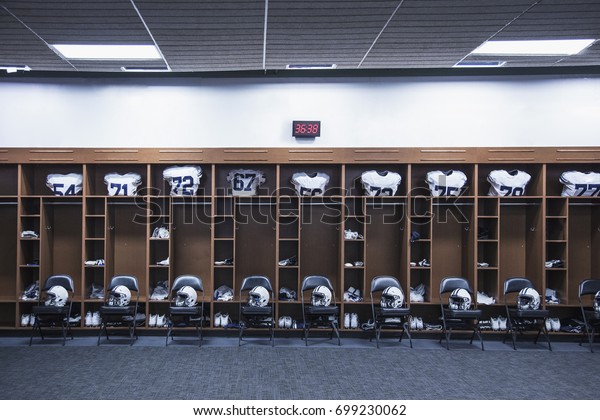 American Football\
locker room in a large stadium. Helmets sitting on chairs and\
jersey`s resting in the\
lockers
