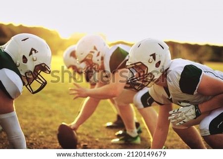 American football players lined up for a scrimmage on a playing field outside during a team practice in the late afternoon