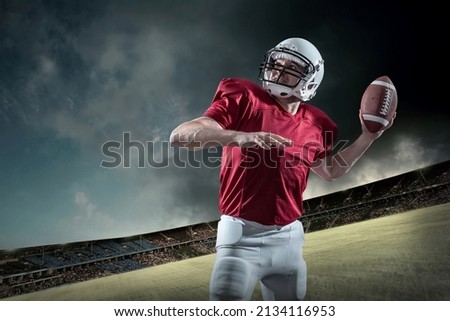 American football sportsman player with ball in action on stadium under lights of background. Sport, proud footballer in white helmet and red shirt ready to play.