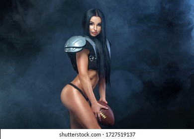 American football. Young sporty brunette wearing sexy uniform of rugby football player posing with ball in smoke isolated on black background