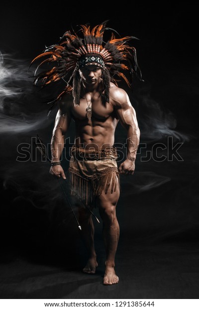 American Indian\
Apache warrior chief  in traditional clothing and feathered\
headdress with weapon. Indian chieftain of the tribe with muscled\
strength body on smoke dark\
background.