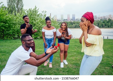 american male standing on one knee asking woman marriage giving ring proposing surprised shocked while gathering group friends all around in shock confusing evening cloudy autumn spring in the park