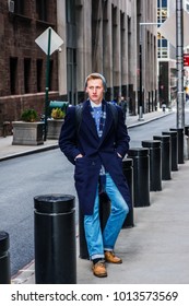 American Man Traveling In New York, Wearing Blue Long Overcoat, Scarf, Jeans, Leather Shoes, Cuffed Knit  Hat, Carrying Back Bag, Hands In Pockets, Sitting On Metal Pillar On Vintage Street, Relaxing.