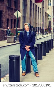 American Man Traveling In New York, Wearing Blue Long Overcoat, Scarf, Jeans, Leather Shoes, Cuffed Knit Hat, Carrying Back Bag, Sitting On Metal Pillar On Vintage Street, Relaxing. 
