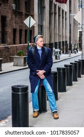 American Man Traveling In New York, Wearing Blue Long Overcoat, Scarf, Jeans, Leather Shoes, Cuffed Knit  Hat, Carrying Back Bag, Sitting On Metal Pillar On Vintage Street, Relaxing. 
