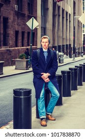 American Man Traveling In New York, Wearing Blue Long Overcoat, Scarf, Jeans, Leather Shoes, Cuffed Knit  Hat, Carrying Back Bag, Sitting On Metal Pillar On Vintage Street, Relaxing. 
