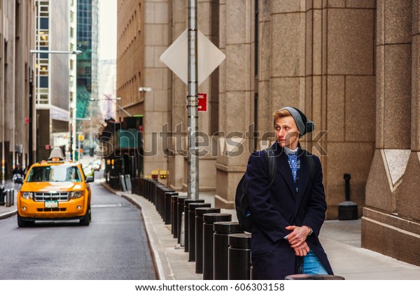 American man traveling in New York, wearing\
blue long overcoat, scarf, cuffed knit beanie hat with pom pom,\
carrying back bag, sitting on metal pillar on vintage street,\
relaxing. Car on\
background.