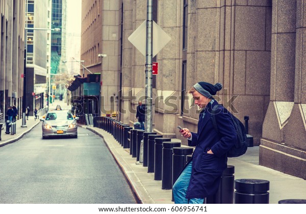 American man traveling in New York, wearing\
blue long overcoat, scarf, cuffed knit beanie hat with pom pom,\
carrying back bag, sitting on vintage street, texting on cell\
phone. Car on\
background.\
